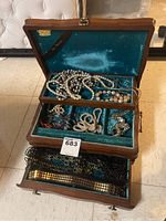 Open wood jewelry box showing teal velvet lining and assorted jewelry inside, including necklaces and belts.