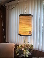 Mid century modern hanging lamp with woven textured cylindrical shade, dark trim, and pull cord, hanging near vertical blinds and a beige chair.