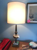 Full view of the table lamp illuminated, showing tall cream fabric lampshade and ornate base on wood surface with red suitcase nearby.
