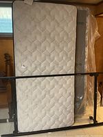 Mattress, boxspring, and metal bed frame stacked upright in a basement setting. Mattress front-facing with quilted pattern visible.
