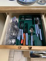 Cutlery drawer with blue handled knives, forks, spoons in green plastic tray and a zip-lock bag of additional metal spoons