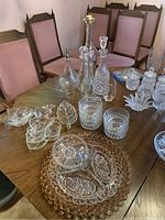 Wide view of table showing multiple crystal decanters, glasses, divided dishes, and bowls arranged alongside wooden dining chairs.