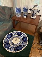 Photo showing a large blue and white decorative wall clock, a tall vase with handles, a pair of bookends with blue landscape pattern, and a small ceramic container on a wooden table.