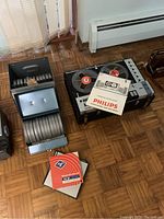 Philips reel-to-reel tape recorder loaded with two reels, instruction manual on top, next to metal storage cabinet and envelope sleeves on parquet floor
