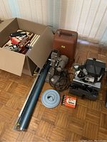 Photo showing a vintage slide projector with a power cord, a brown carrying case, two blue cylindrical projector screens, a small boxed slide viewer, a slide maker box, a large slide viewer device, a metal slide reel, and a cardboard box filled with slides and reels.