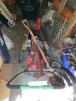 Photo shows electric lawn mower loaded with multiple wooden handled yard hand tools including shovels, rakes, and garden forks.