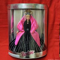Full front view of Barbie doll in original cylindrical packaging, showcasing the black dress with silver streaks and pink wrap