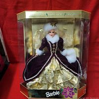 Front view of the Barbie doll in original sealed packaging, showing the purple velvet coat and gold skirt.