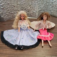 Two Barbie dolls seated on a wooden surface with a silver textured background; one in a lavender skirt with floral pattern and white lace top, the other in a pink skirt with cream shawl.