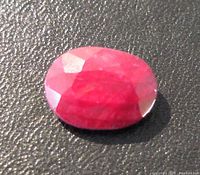 Close-up photo of the oval faceted ruby gemstone on a black textured background showing deep red color and facets.