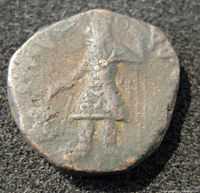 Obverse side of ancient Kushan Kanishka coin showing a standing figure in traditional garb, exhibiting wear and patina from age