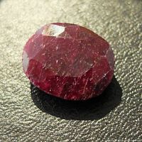 Close-up photo of oval faceted ruby gemstone showing deep red color with natural inclusions and surface texture.