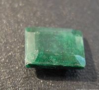 Close-up photo of the rectangular deep green emerald gemstone on a black surface, showing facet cuts and natural inclusions.