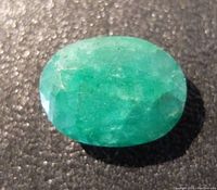 Close-up photo of oval-shaped green emerald gemstone showing faceting and natural inclusions on black background.