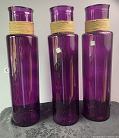 Photo showing three tall purple glass vases with twine wrapped around their necks, standing side by side on a patterned black surface against a white background.