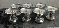 Eight silver Debi Lilly Manhattan candle holders arranged in two rows on black fabric with an ornate damask pattern, showing the front and side profiles.