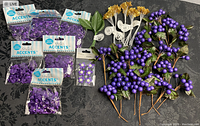 Top view of unopened packs of loose purple flower petals, small artificial white flower stems with tags, packages of tiny purple flower accents, and several artificial purple berry branches with green leaves arranged on a black patterned surface.