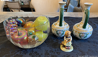 Glass bowl filled with multiple colorful iridescent glass fruits alongside two ceramic vases with green handles and a small Goebel figurine of a girl holding a book on a black tablecloth.