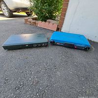 Two Coleman portable camping stoves with closed lids, resting on asphalt near a brick wall and garden bed.