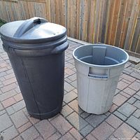 Two large plastic garbage pails on outdoor paving with wooden fence background, black one has lid, gray one is open
