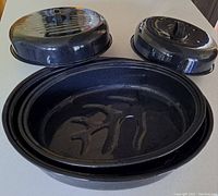Set of two black speckled enamel roasting pans with lids on a table. One oval pan in front and the round pan behind.
