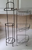 Shows a black metal toilet paper holder with a top horizontal bar and circular base designed for extra rolls alongside a silver metal wire corner shelf with three baskets.