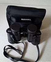 Front view of Bushnell Falcon binoculars with lens caps on, neck strap and carrying case.