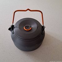 Top view photo of a lightweight portable camping kettle with black body and orange handle and lid knob on a plain surface.