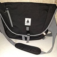 Front view of black and gray messenger laptop carrier bag with shoulder strap and front buckle, showing logo patch and zippered pocket.