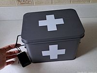 Closed view of the black metal medical organizer box with white cross symbol on top and side. Hand holding the box handle.