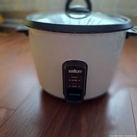 Full frontal view of the white Salton rice cooker on a wooden floor showing the glass lid with a black handle and the front control panel.