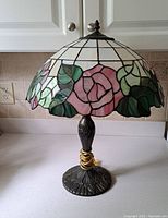 Full view of Tiffany style stained glass lamp showing pink rose and green leaf pattern on shade and antique bronze base.