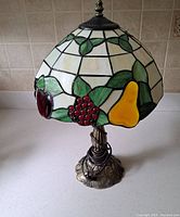 View of Tiffany style stained glass lamp showing fruit motifs including yellow pear, cluster of red berries, and apple on white background.