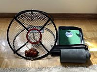 Golf practice net with central red hoop net, white netting, and bag of orange golf balls alongside a green putting putting green mat with single hole and black tray.