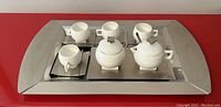Full view of the tea service set arranged on the large stainless steel tray including the cups, saucers, cream and sugar bowl.