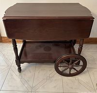 Front view of vintage wooden drop leaf trolley tea service cart with drop leaf sides down, showing large spoked wooden wheels and lower shelf.