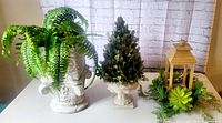 Photo showing three artificial decorative pieces: an artificial fern in a white ornate pot, a leafy cone shaped topiary, and a wooden lantern with succulents around base.