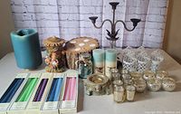 Various candles, candle holders, candelabra, taper candles, and tealight holders arranged on a flat surface against a brick wall background.