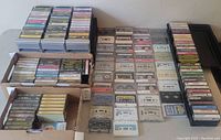 Several hundred cassette tapes neatly arranged with some in plastic storage boxes and others in cardboard boxes, showcasing the variety and quantity of tapes included.