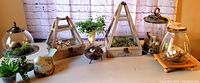Handmade nature decor items including several glass jars with metal and wooden lids, pyramid-shaped terrariums, and artificial plants arranged on a table with a brick backdrop.