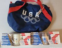 Blue USA Olympic tote bag with red straps and 2 boxed plastic cups from 1984 LA Games, displayed on table.