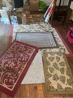 Five rugs displayed on a wooden floor with visible wear and a finger pointing to a furniture indentation on one rug.