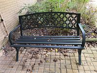 Full view of outdoor metal bench showing lattice backrest, slatted seat, and curved armrests on a paved surface with surrounding garden.