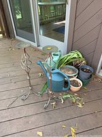 Two metal candleholders with twisted design, tripod feet, circular tops, accompanied by an assortment of plastic garden pots and two watering cans, photographed on a wooden deck near glass door.