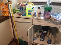 Photo of multiple cleaning bottles and containers including Arm & Hammer Baking Soda bag on left, various spray bottles, large yellow and white jug, Miracle Gro Flower Food, Bona cleaners, and other assorted cleaning products on kitchen counter and inside under-sink cabinet.