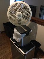 White Lasko oscillating stand fan and five trash cans, one small stainless steel step-on can and four black plastic cans with visible damage on one.
