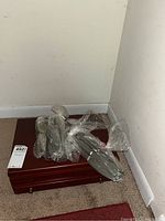 Photo showing wooden storage box and multiple plastic-wrapped bundles of silver-colored flatware items on carpet.