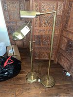 Two brass floor lamps with adjustable arms and rectangular shades placed against a carved wooden screen on a wood floor, showing signs of wear and patina.