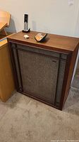 Single vintage Wharfedale floor speaker shown upright with dark wood frame and fabric mesh front, placed beside wooden furniture and carpeted floor.