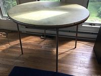 Full view of round folding table with metal folding legs on hardwood floor near window